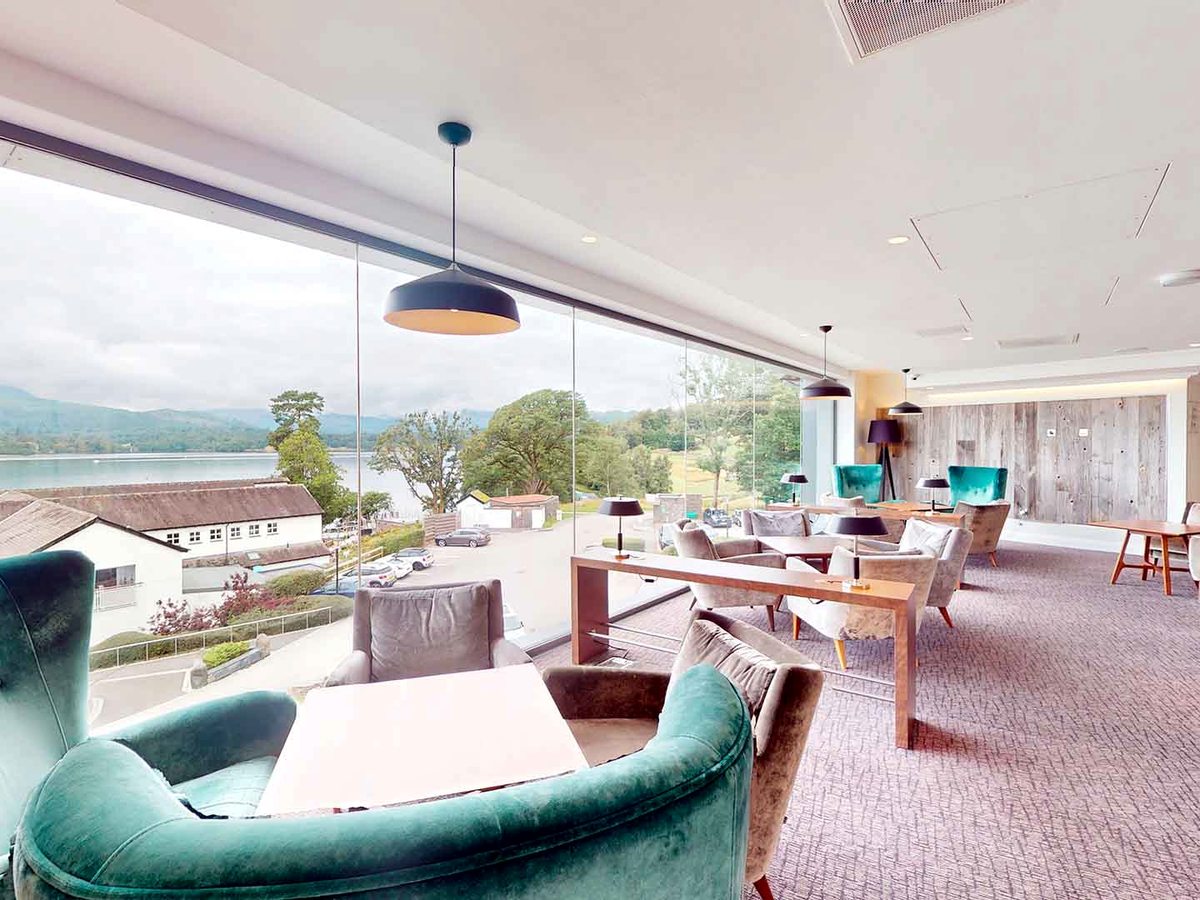 A lounge with teal and grey armchairs around low tables, a wood-panelled wall and a full-height glass wall onto the lake and the fells.