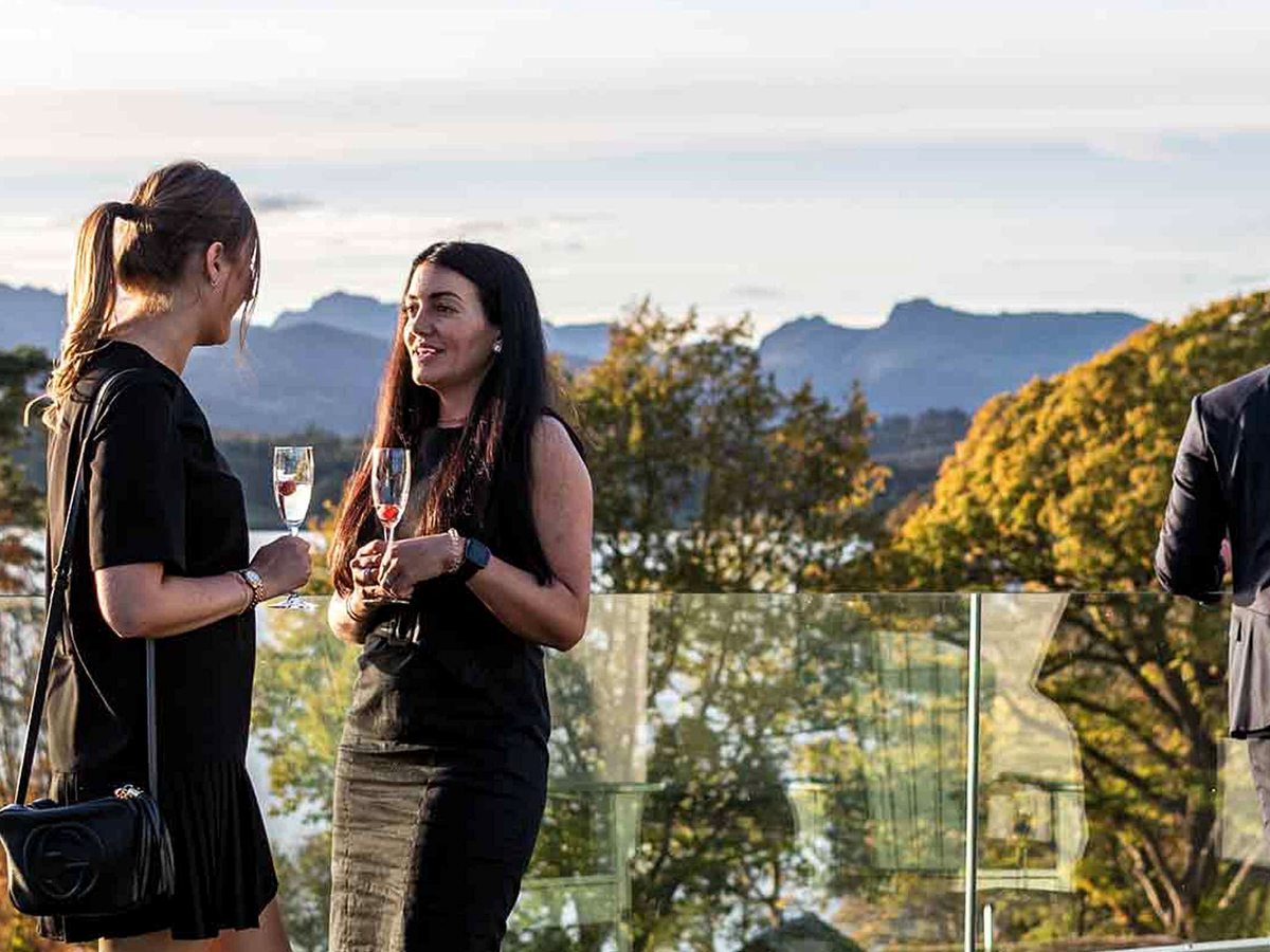 Two guests standing with champagne glasses at the glass balustrade of a terrace, with autumn trees and the fells behind them.