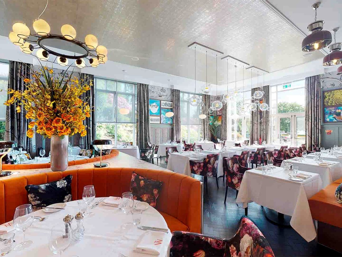 A dining room with curved orange banquettes, floral-print armchairs, white-linen tables, a ring chandelier and a tall vase of yellow flowers.