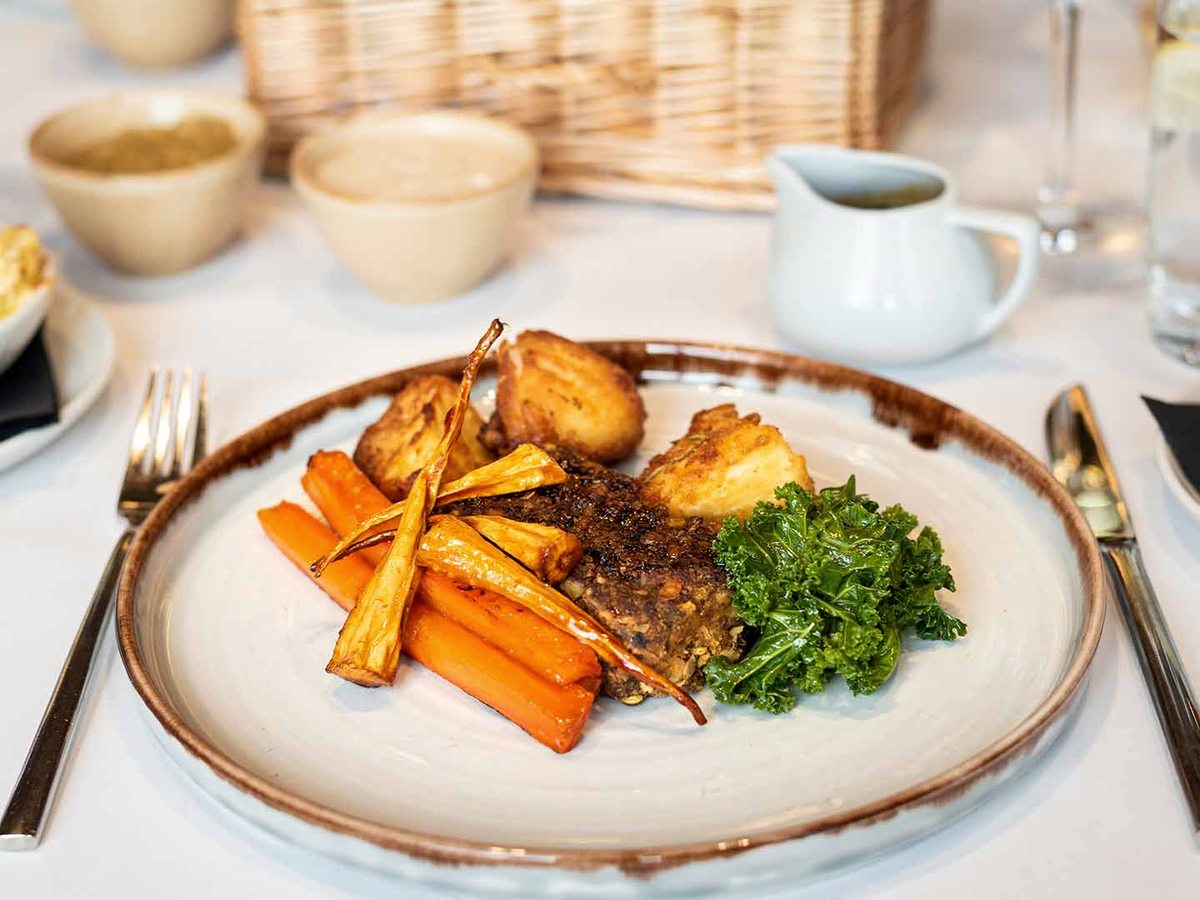 A roast dinner on a rimmed stoneware plate: a wedge of nut roast with roast potatoes, glazed carrots, parsnips and kale, with a jug of gravy and bowls of vegetables alongside.