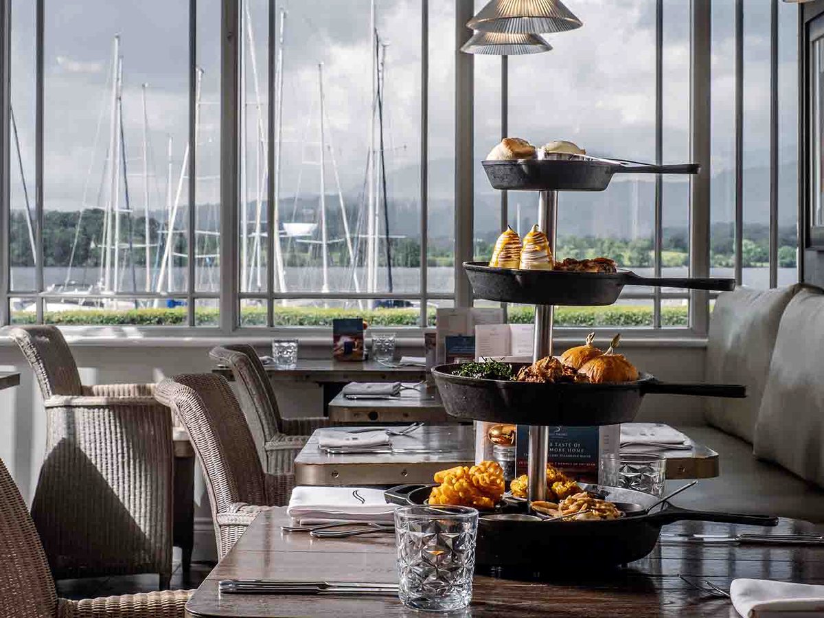 A tiered stand of black cast-iron pans holding buns, savoury bites and pastries on a dark wooden table, with the masts of moored yachts through the window behind.