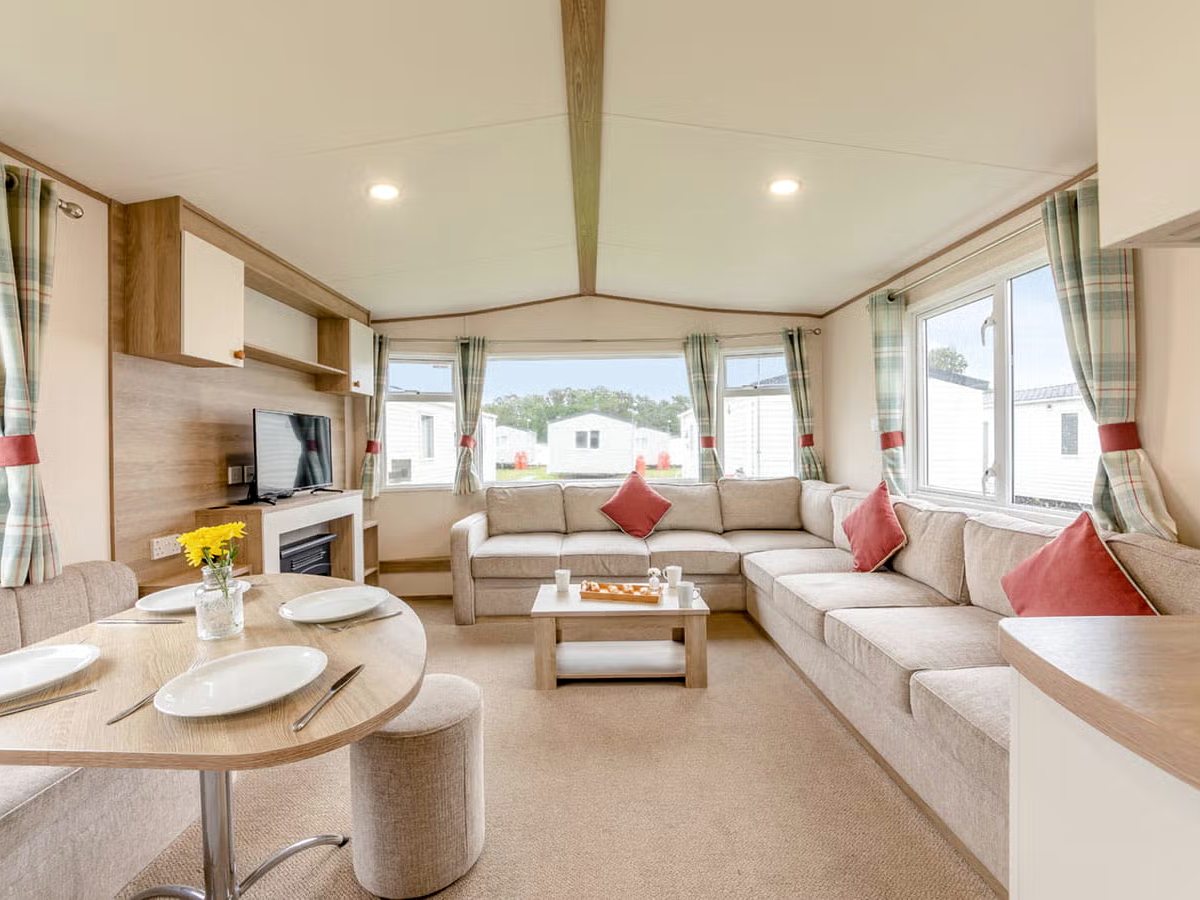 An open-plan caravan lounge with a cream corner sofa and red cushions, a low coffee table, a television above an electric fire and a round dining table with stools by the windows.
