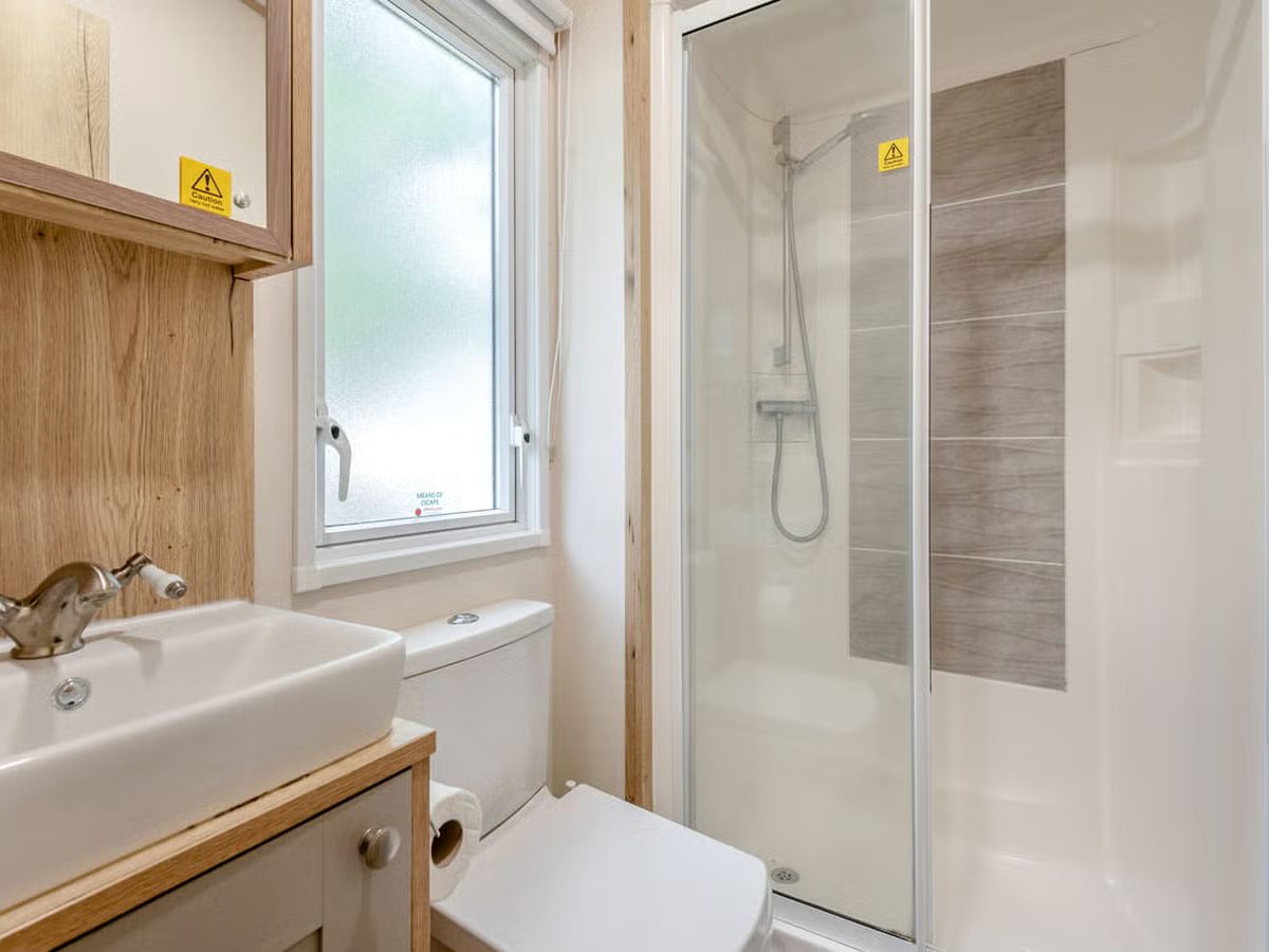 A compact caravan shower room with a sliding-door shower enclosure, a white basin on a wood-effect vanity unit, a toilet and a frosted window.