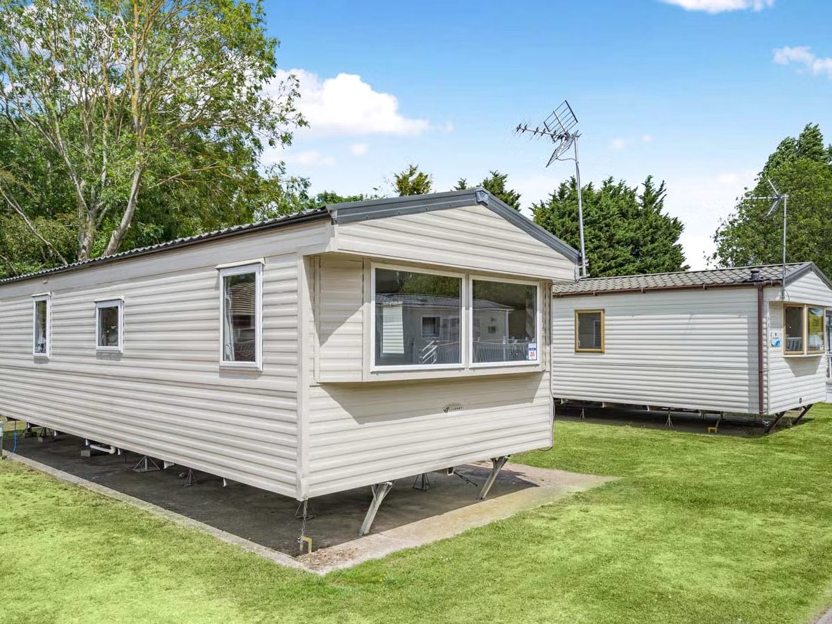 Two cream static caravans standing on mown grass with mature trees behind them and a blue sky with light cloud.