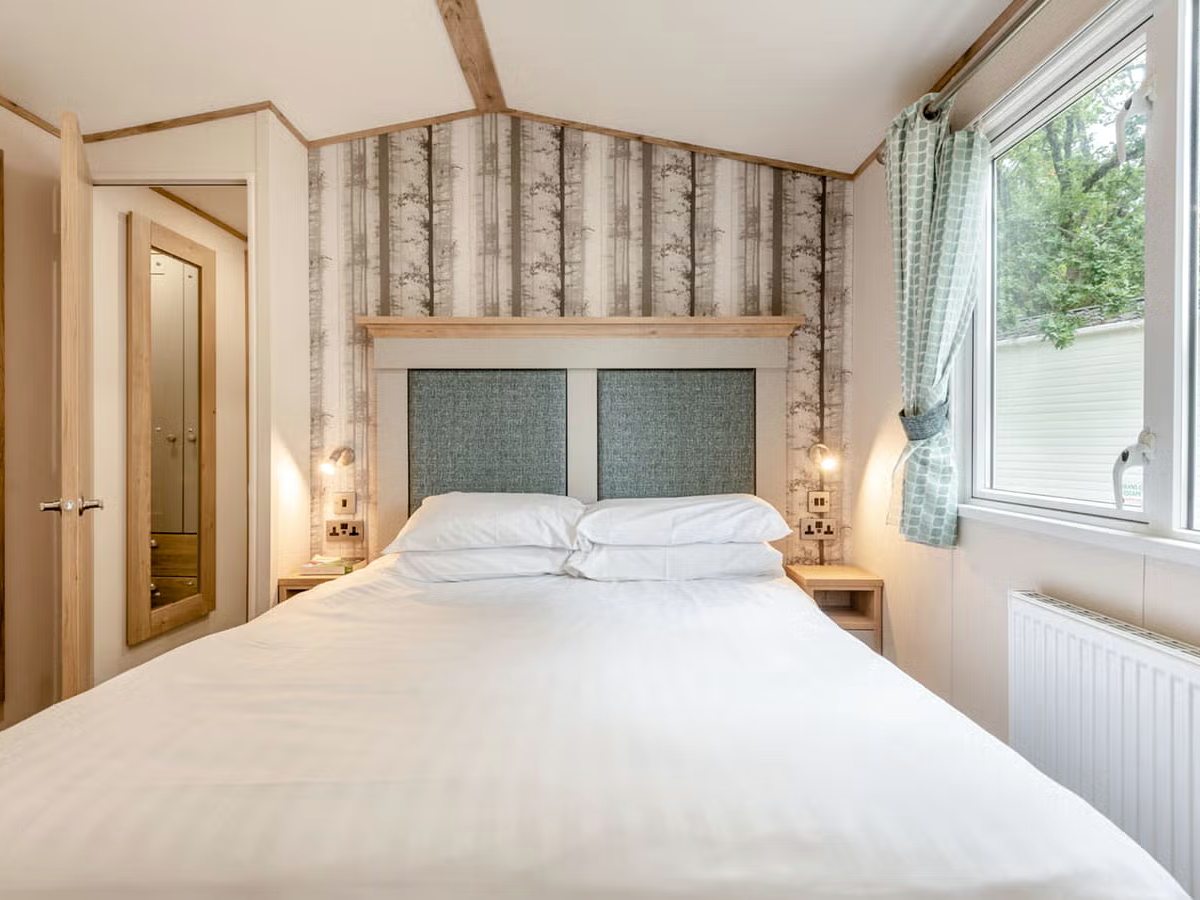 A caravan double bedroom with white bed linen, a padded green headboard, birch-tree wallpaper, small bedside lamps and a window on to trees.
