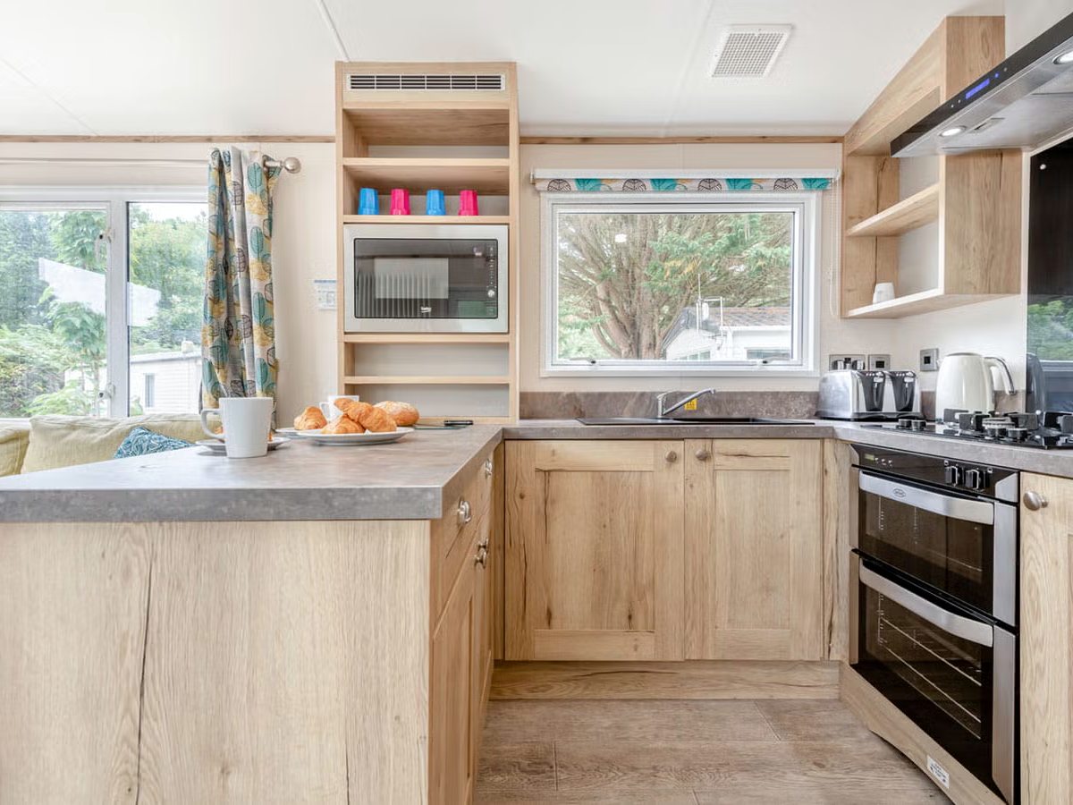 A caravan kitchen in pale oak with a peninsula counter, a built-in microwave, a double oven, a gas hob and a window above the sink.