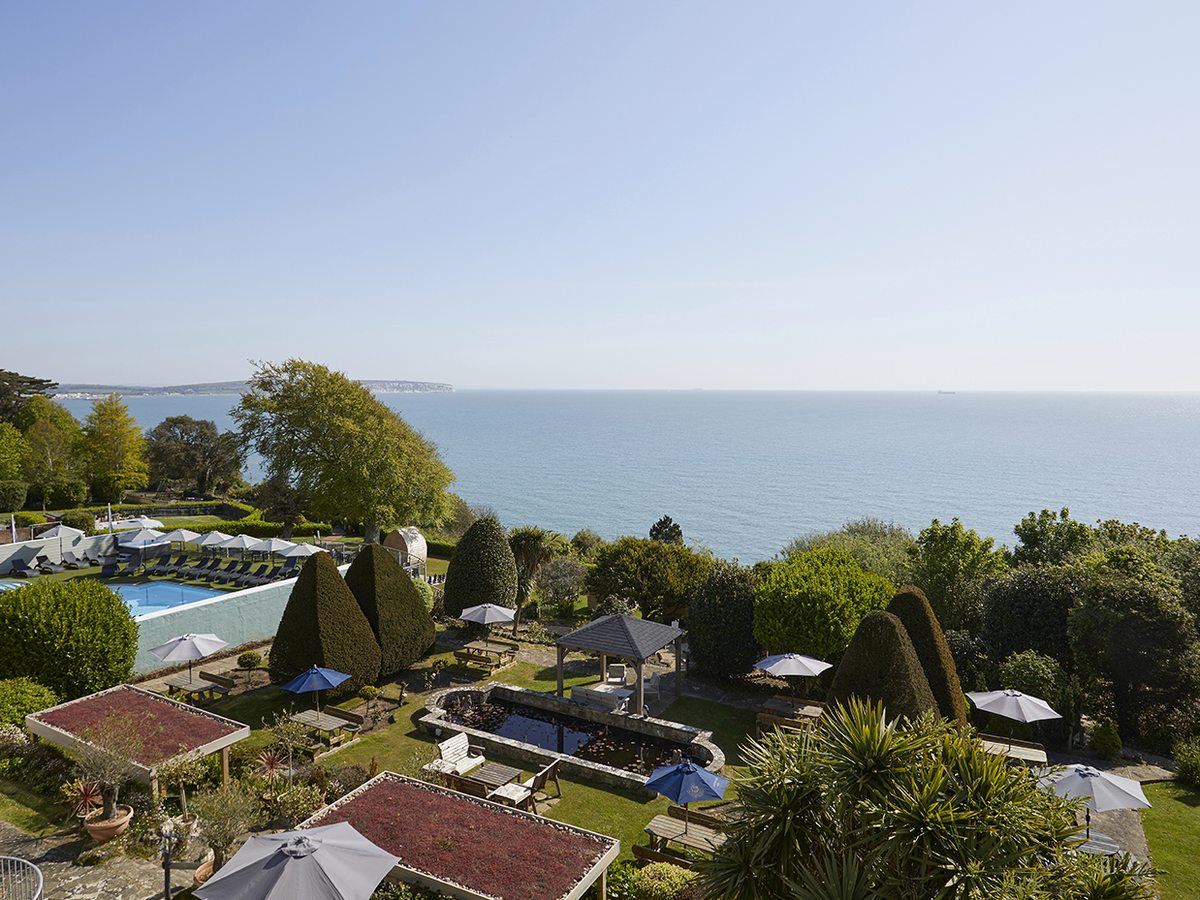 Terraced hotel gardens with clipped topiary, parasols and a pond, an outdoor pool at the far end and the sea and a headland beyond.