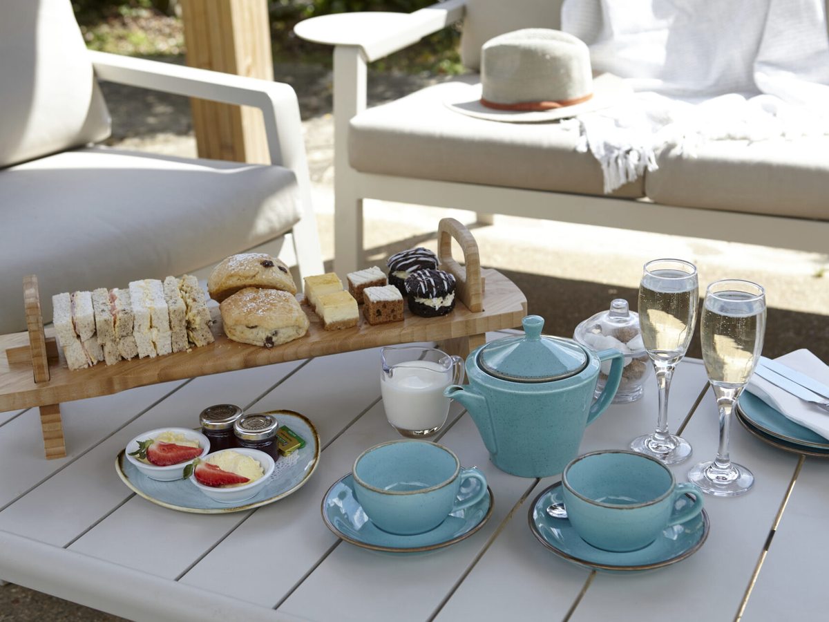 Sandwiches, scones and small cakes on a wooden stand on an outdoor table, with teapots, cups and glasses of sparkling wine.