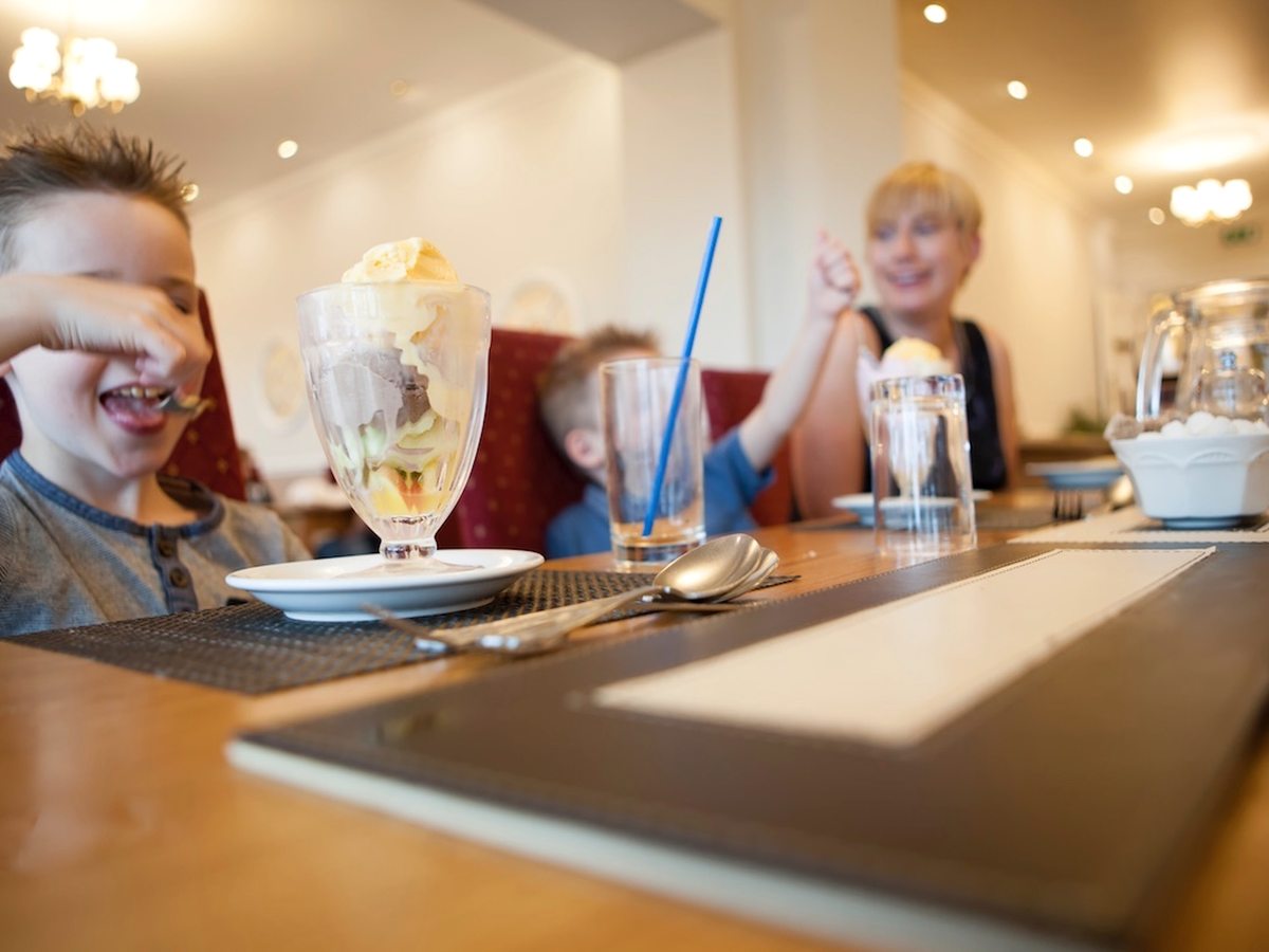 A boy at a restaurant table grinning over a tall ice cream sundae in a glass dish, with a woman and a second child behind him.
