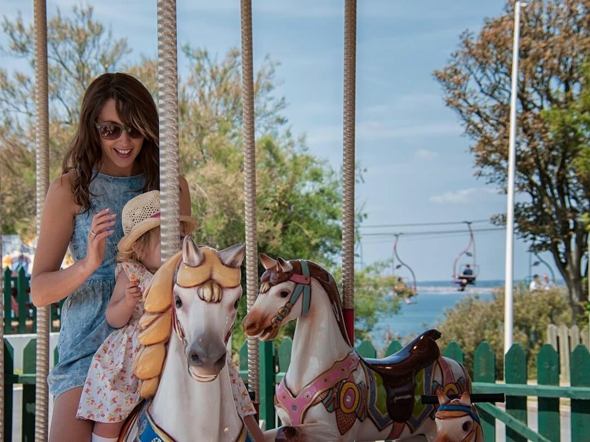 A woman standing beside a small child on a painted carousel horse, with a chairlift and the sea in the background.