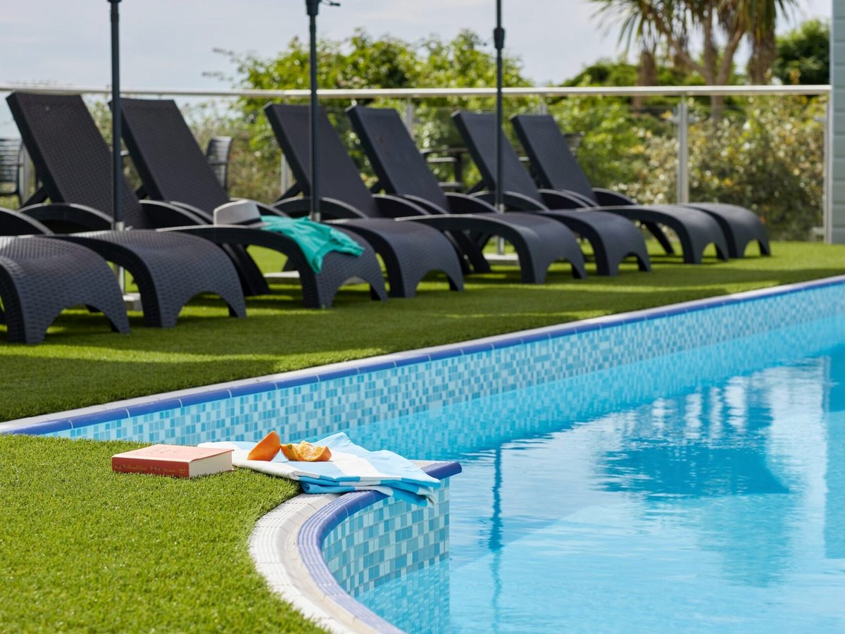 A curved outdoor pool with black sun loungers on artificial grass and towels laid out beside it.