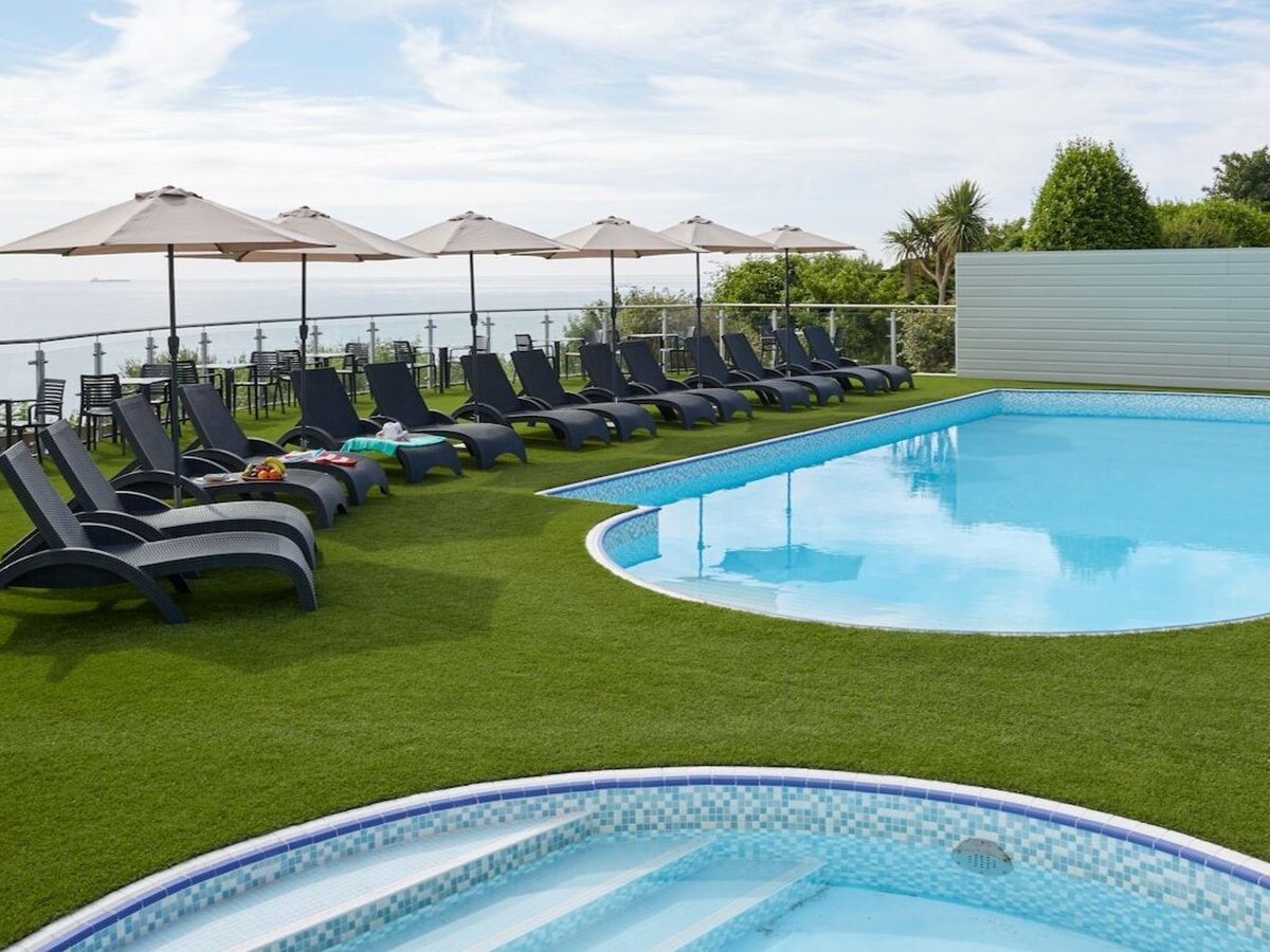 An outdoor pool with a rounded end, sun loungers and parasols in a row along the sea side, a smaller round pool in the foreground and the sea beyond.