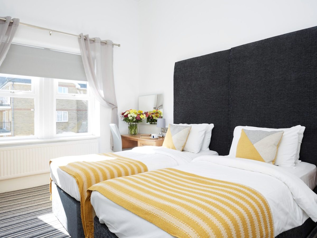 Two single beds with white bedding and mustard throws against a dark headboard, with a window and flowers between them.