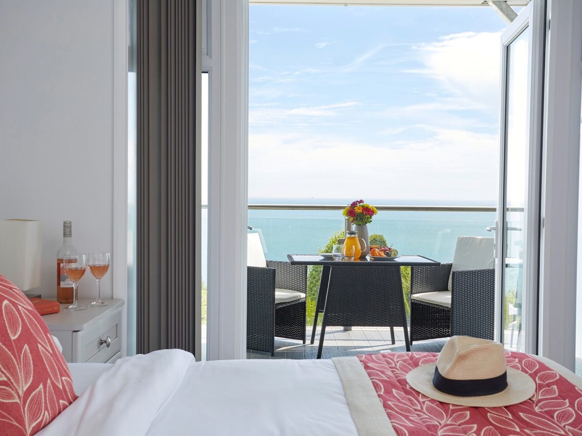 A bed with a coral runner and a straw hat, folding doors open onto a balcony set with a table and chairs above the sea.