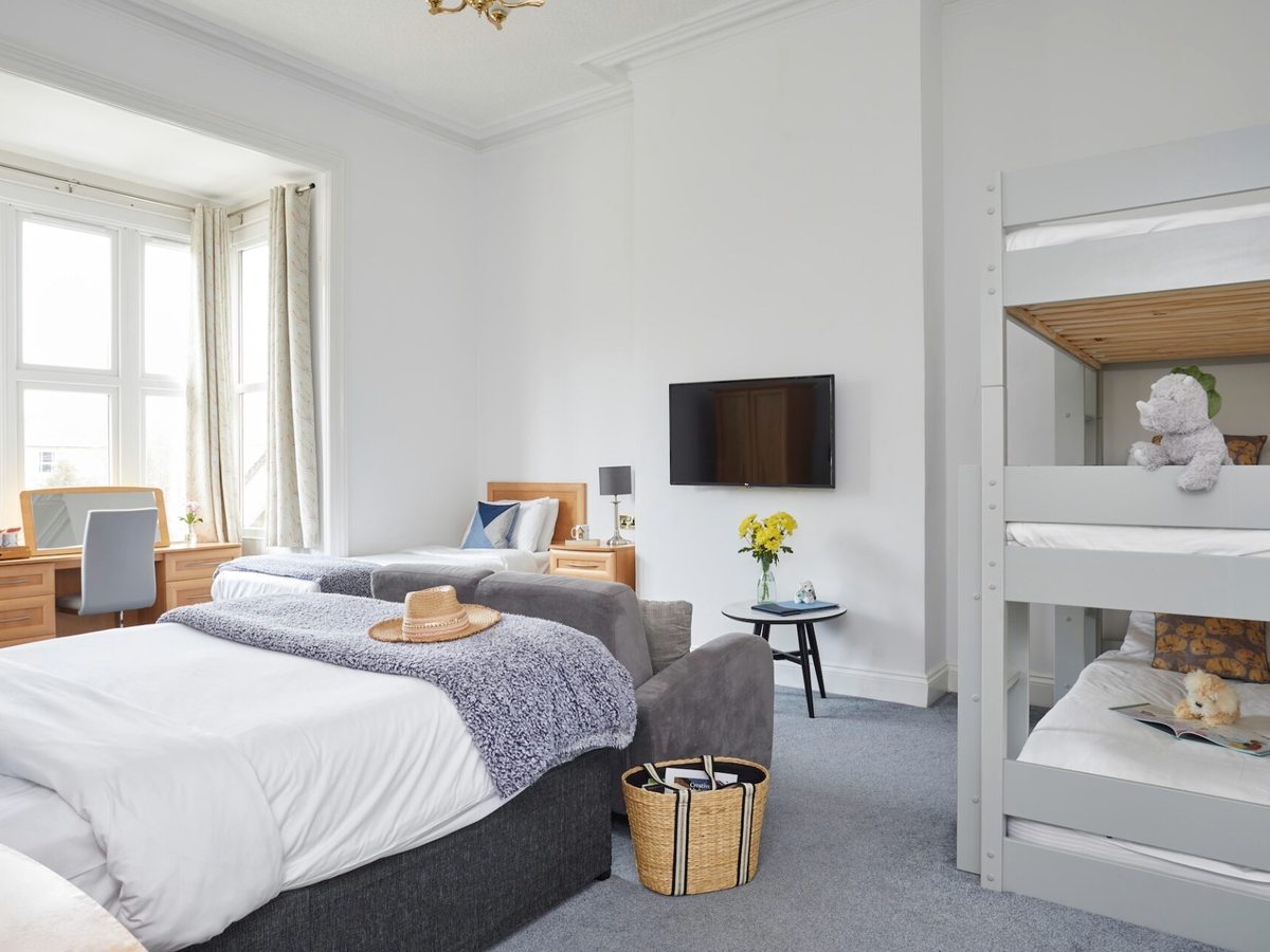 A bright bedroom with a double bed, a single bed, a grey sofa and white bunk beds, with a bay window and a desk.