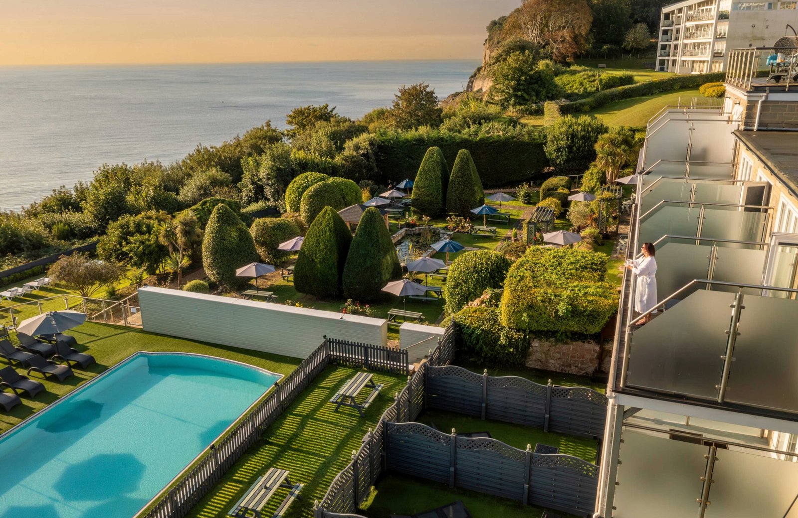 The clifftop gardens at Luccombe Hall Hotel above Shanklin, with the outdoor pool and sun loungers, terraced lawns and topiary, a guest wing to one side and Sandown Bay beyond.