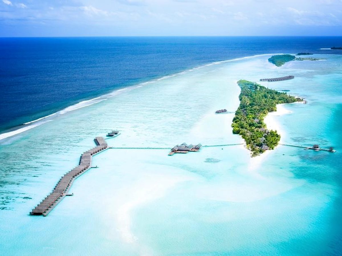 Aerial view of a long narrow palm-covered island with a line of overwater villas running out along a jetty over a pale turquoise lagoon and reef.