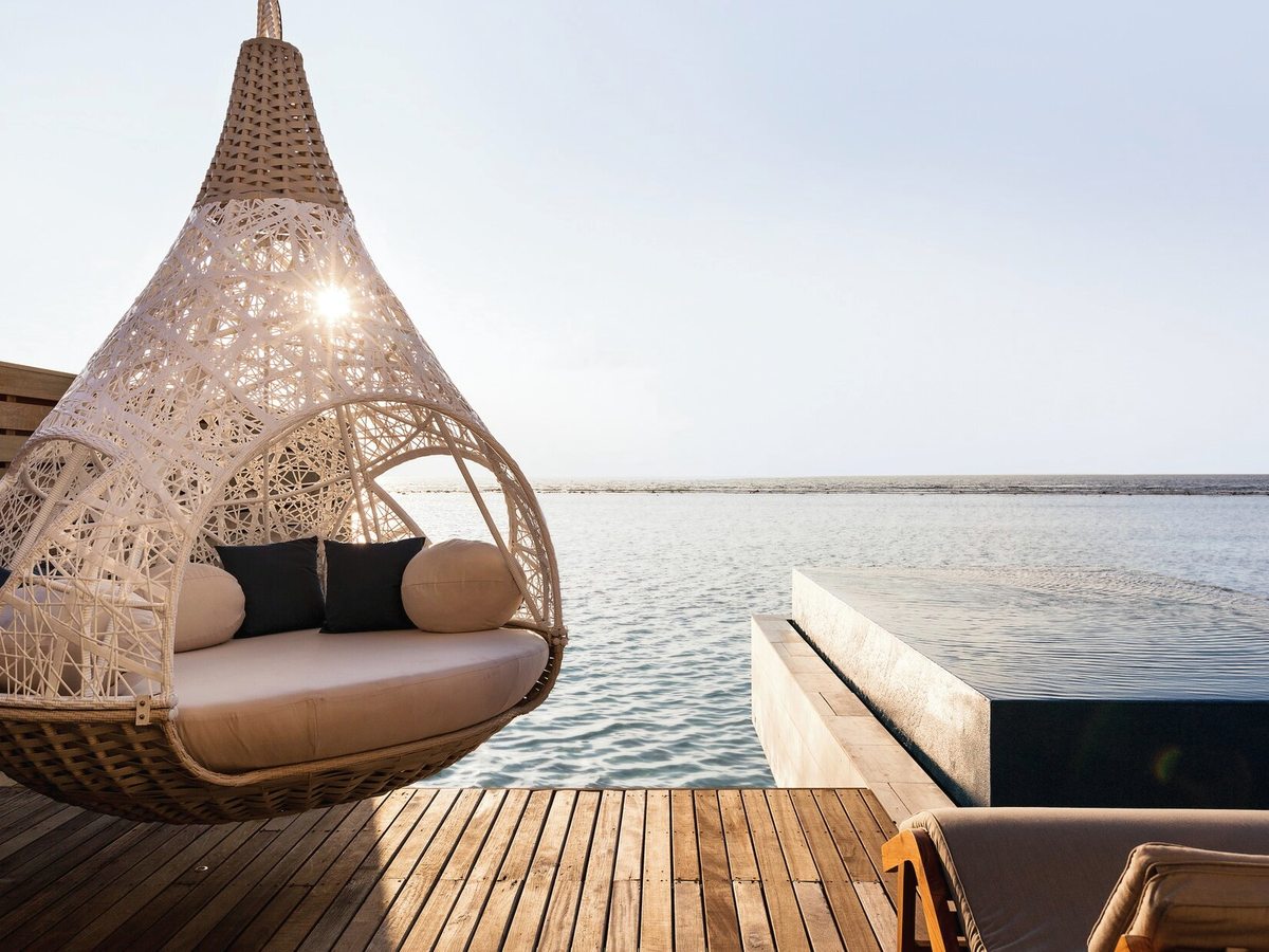 A hanging woven daybed on a timber deck beside a small infinity pool, with calm lagoon water and a distant sandbank beyond.