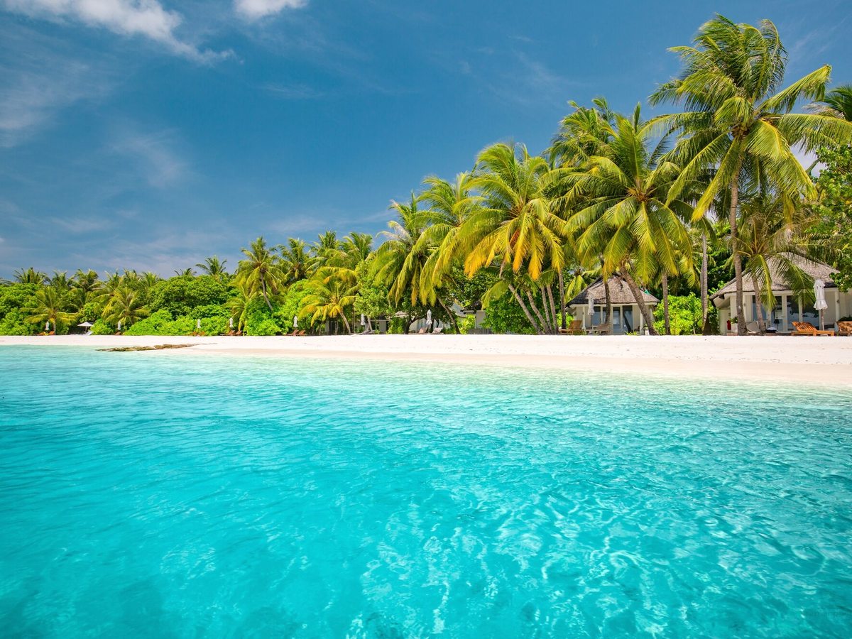Clear turquoise shallows in front of a white sand beach backed by coconut palms and low thatched beach villas.