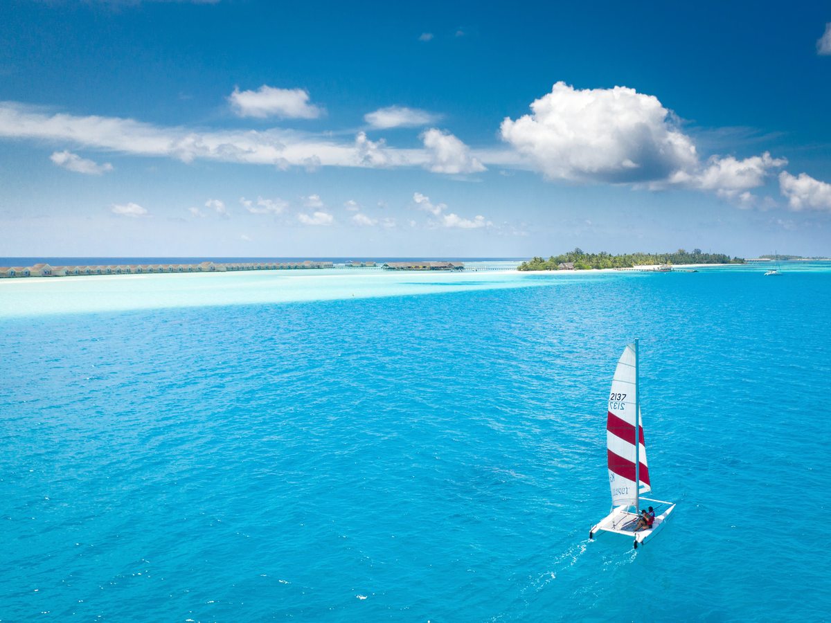 A small catamaran with a red and white sail crossing deep blue water, with the island and its overwater villas along the horizon.