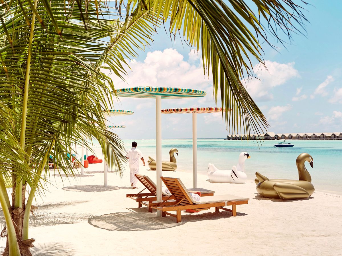 Sun loungers and striped parasols under coconut palms on white sand, with inflatable swans on the shore and overwater villas across the turquoise water.
