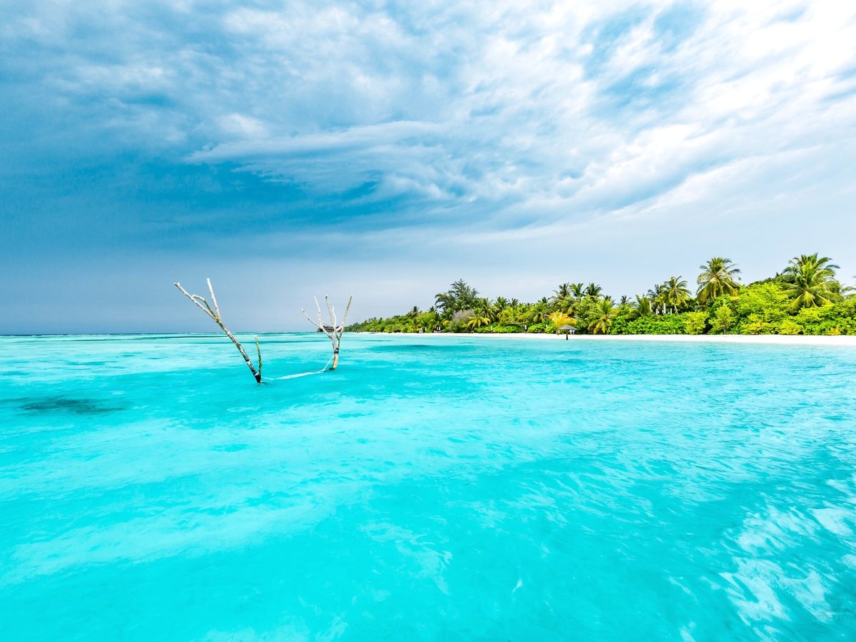 Bright turquoise shallow water in the foreground with the palm-covered island and its white beach along the horizon under a cloudy sky.