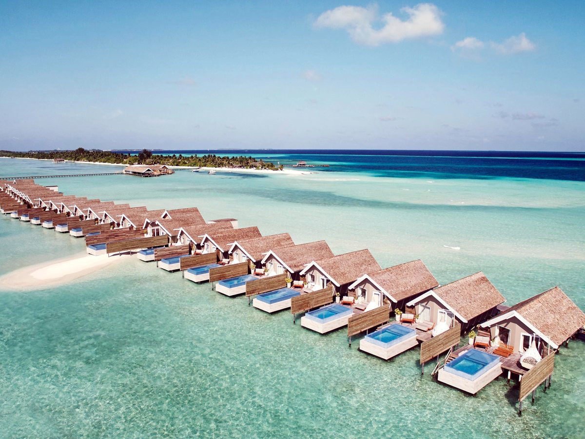 Aerial view of thatched overwater villas in a curving line, each with a small blue plunge pool on its deck above pale turquoise water.