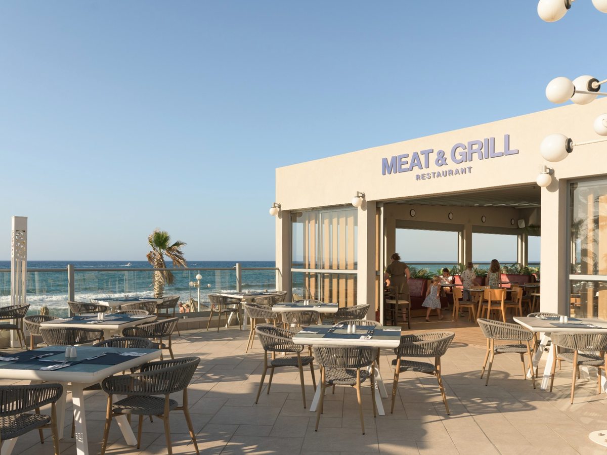 An open-sided restaurant signed Meat & Grill, with wicker chairs and laid tables on a stone terrace beside the sea.