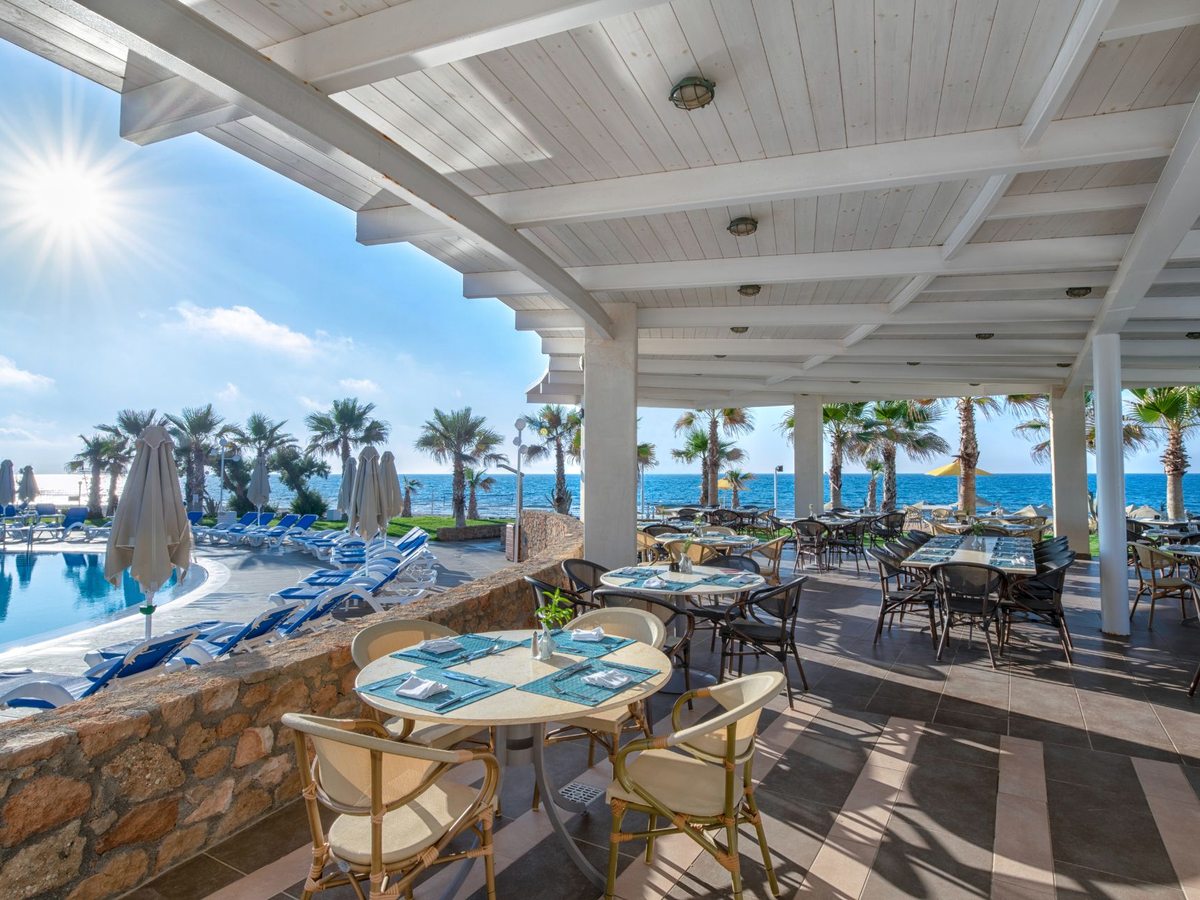 A covered restaurant terrace with laid tables and wicker chairs, looking over pool loungers to the sea.