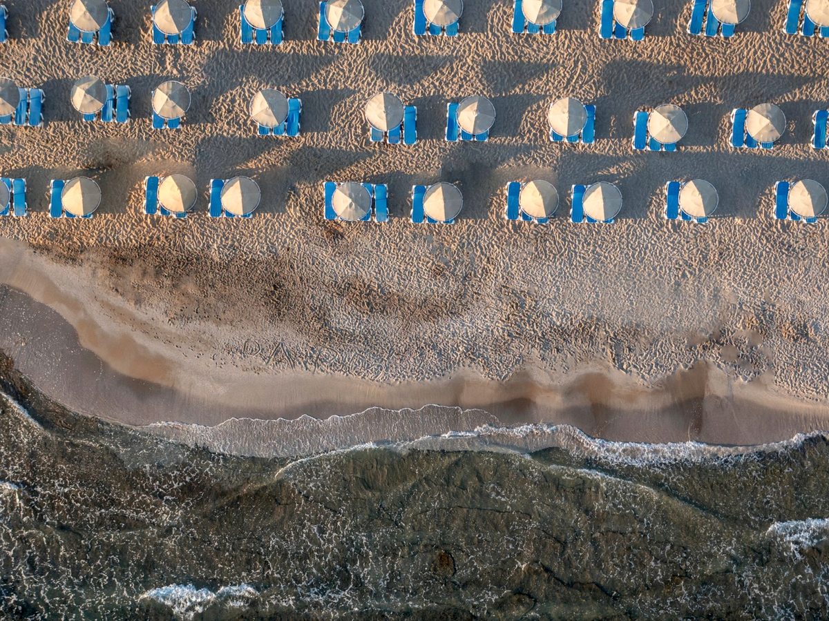 An overhead view of rows of blue sunbeds and parasols on pale sand, with waves breaking along the shore.