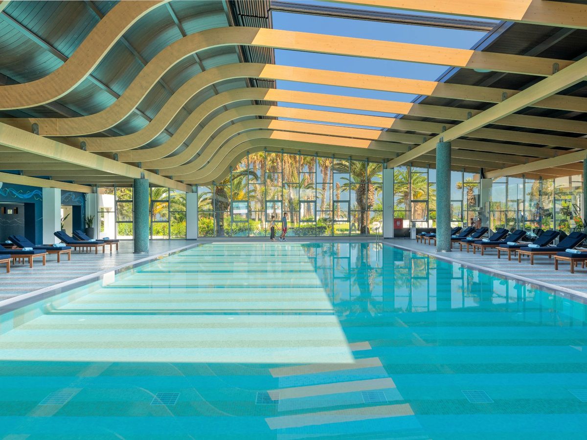 A large indoor pool under a wave-shaped timber ceiling, with sun loungers along the glazed wall.