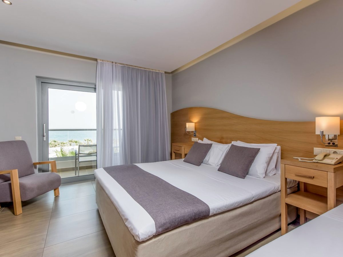 A double bed with a curved wooden headboard and a grey runner, an armchair beside the balcony doors and the sea beyond.