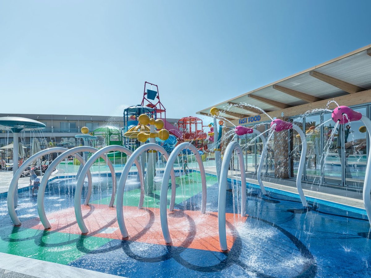A shallow splash area with metal spray arches and water jets, with the slides and a snack counter behind.