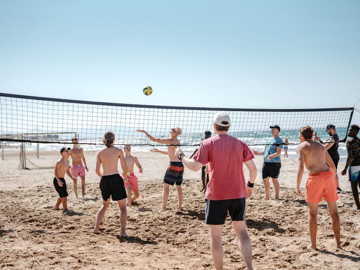 A group of players reaching for a volleyball over a net on the sand, with the sea behind.