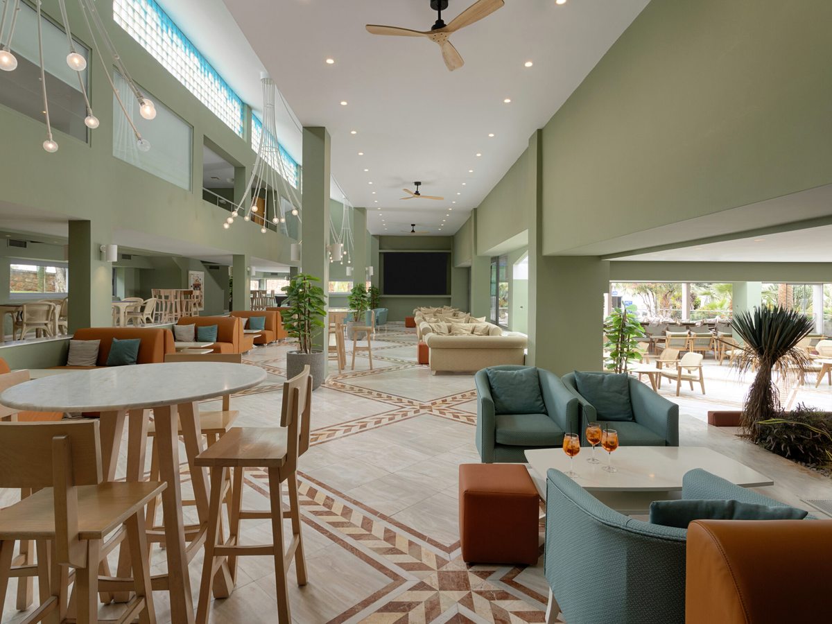A bright open lobby lounge with green and tan sofas, high tables and stools under pendant lights, opening onto a terrace.