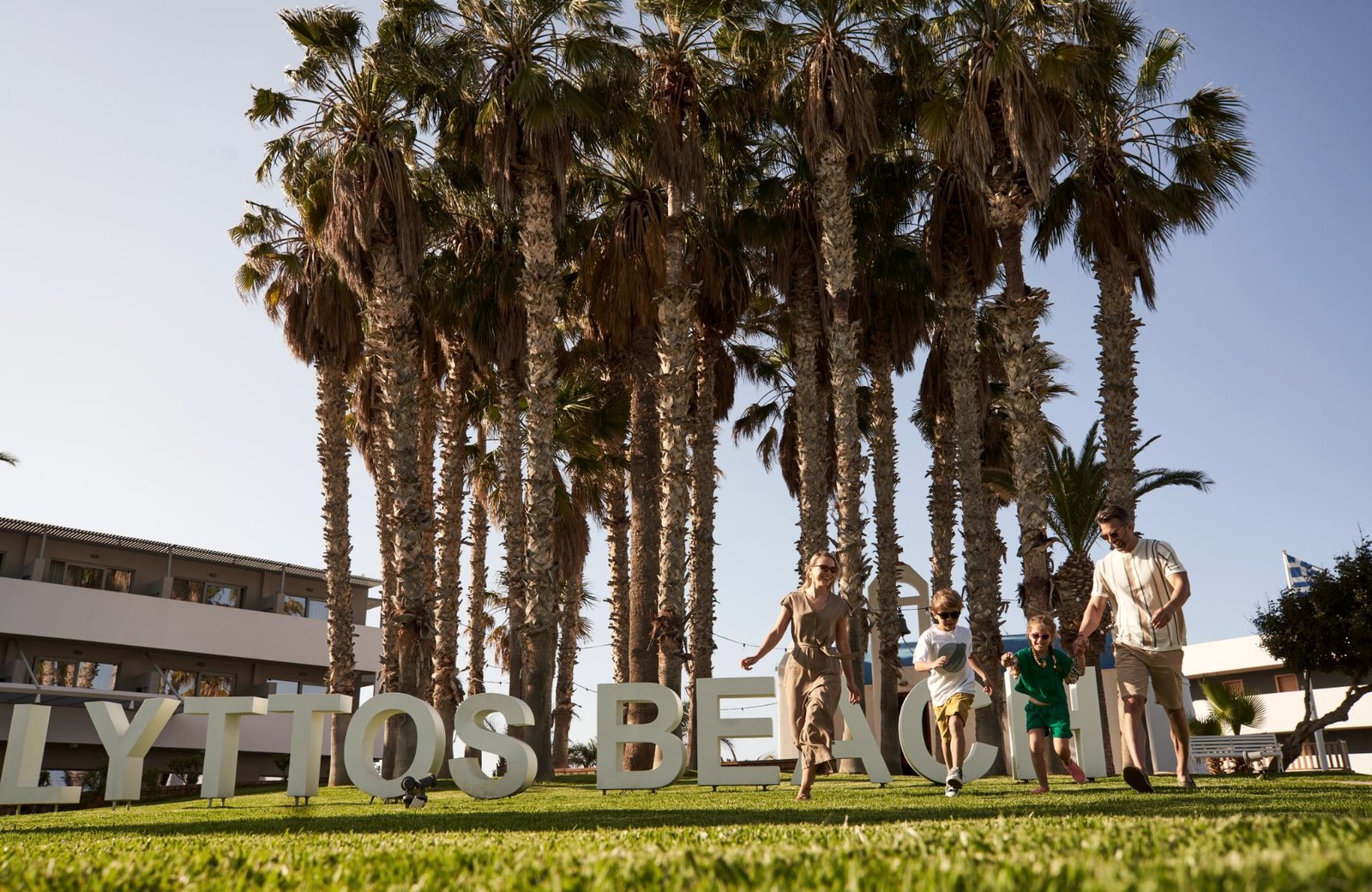 A family walking across a lawn past tall white letters spelling Lyttos Beach, with a row of palm trees and low white resort buildings behind.
