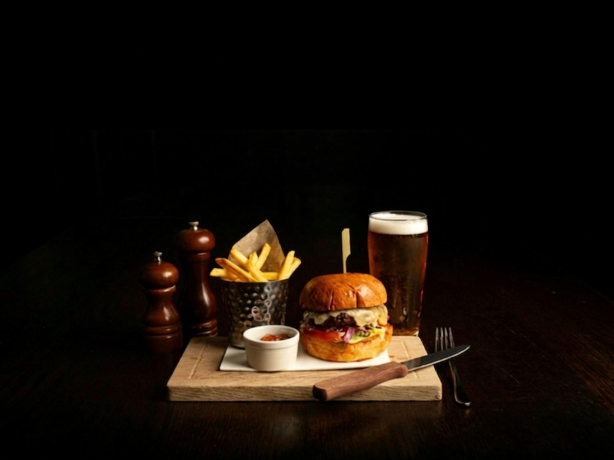 A burger in a brioche bun with a metal cup of chips, a dip and a pint of beer on a wooden serving board.