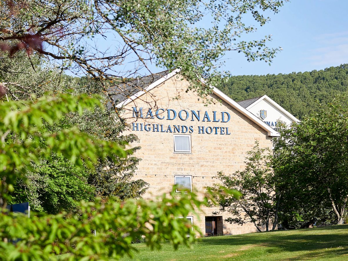 A sandstone hotel gable with a large sign reading Macdonald Highlands Hotel, framed by green leaves, with lawn and pine woods around it.