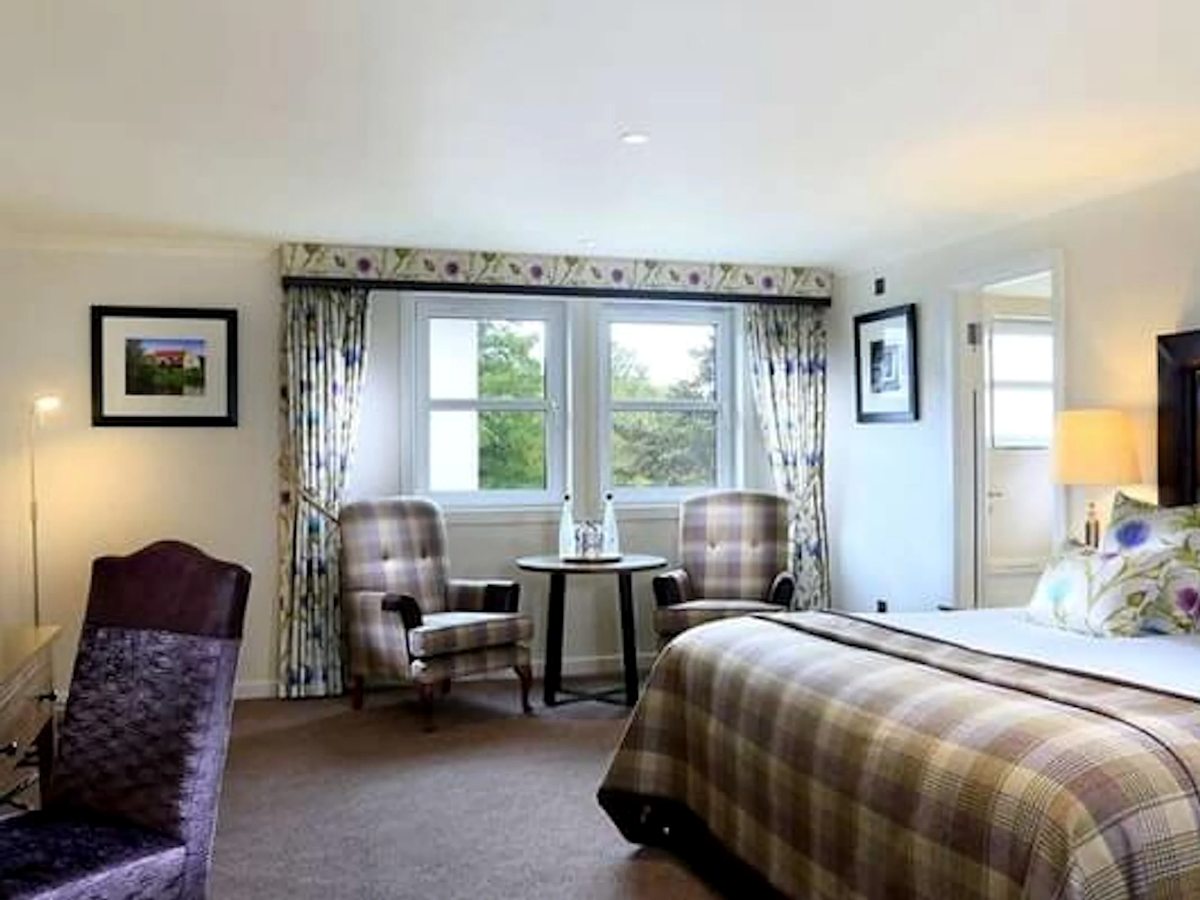 A hotel bedroom with a tartan bedspread, two tartan armchairs and a small round table by a bay window with floral curtains.