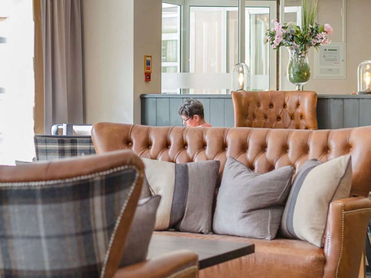 A hotel lounge with a buttoned tan leather sofa and armchairs, tartan and cream cushions, a low table and a vase of flowers by a bright window.