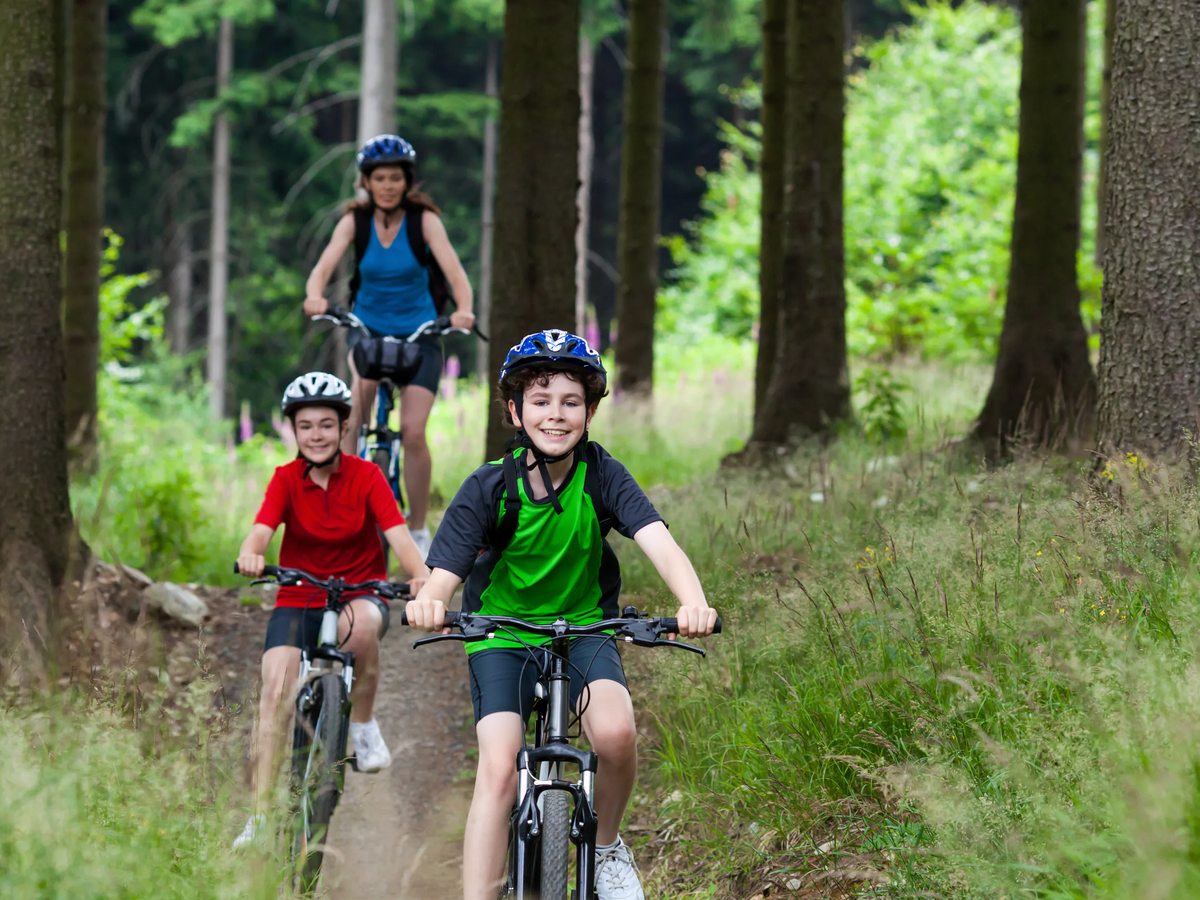 Two children in helmets riding bikes along a sandy forest trail with an adult following, tall pines and long grass on either side.