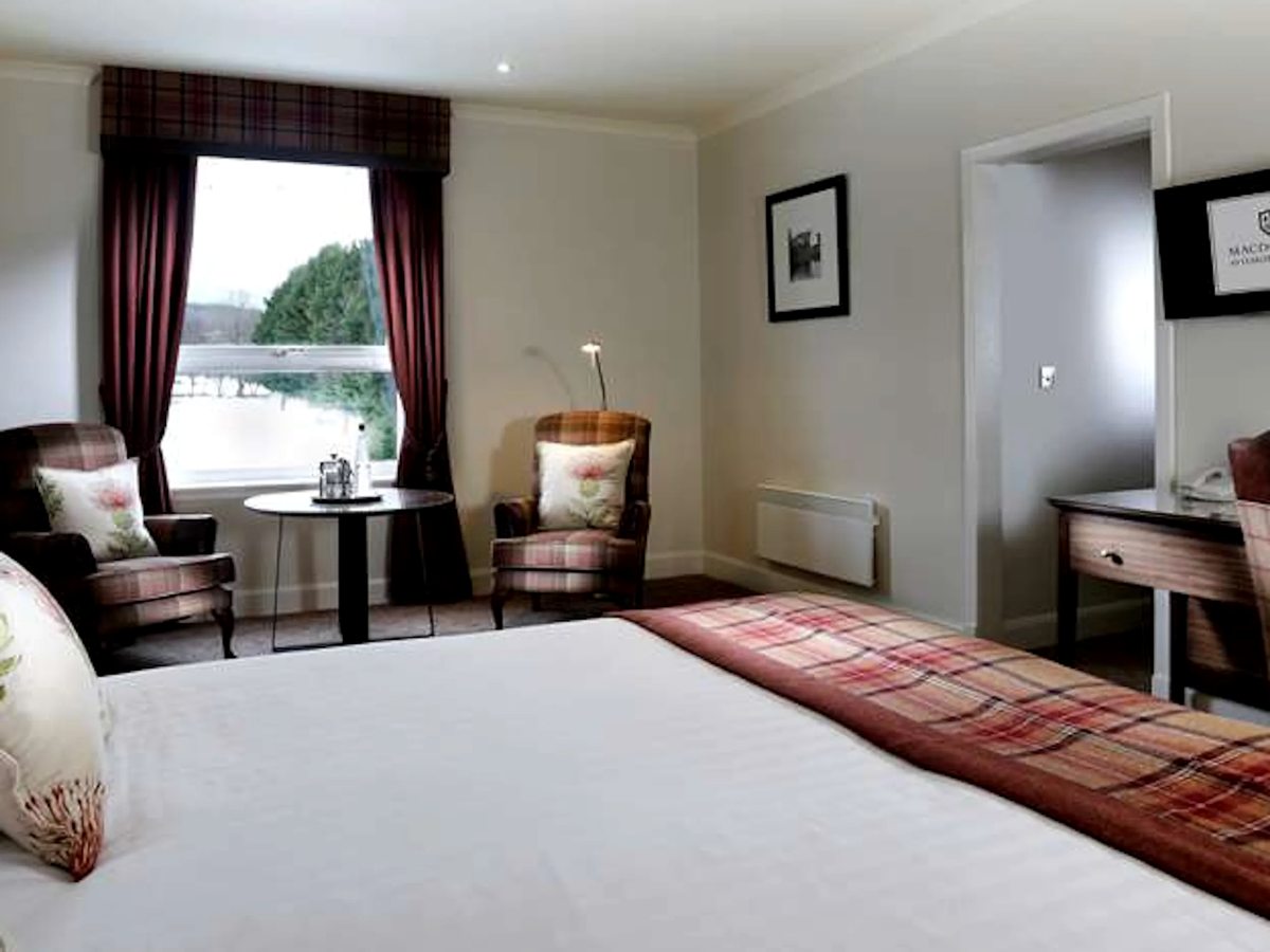 A hotel bedroom with a red tartan runner across a white bed, two armchairs, a writing desk and a curtained window.