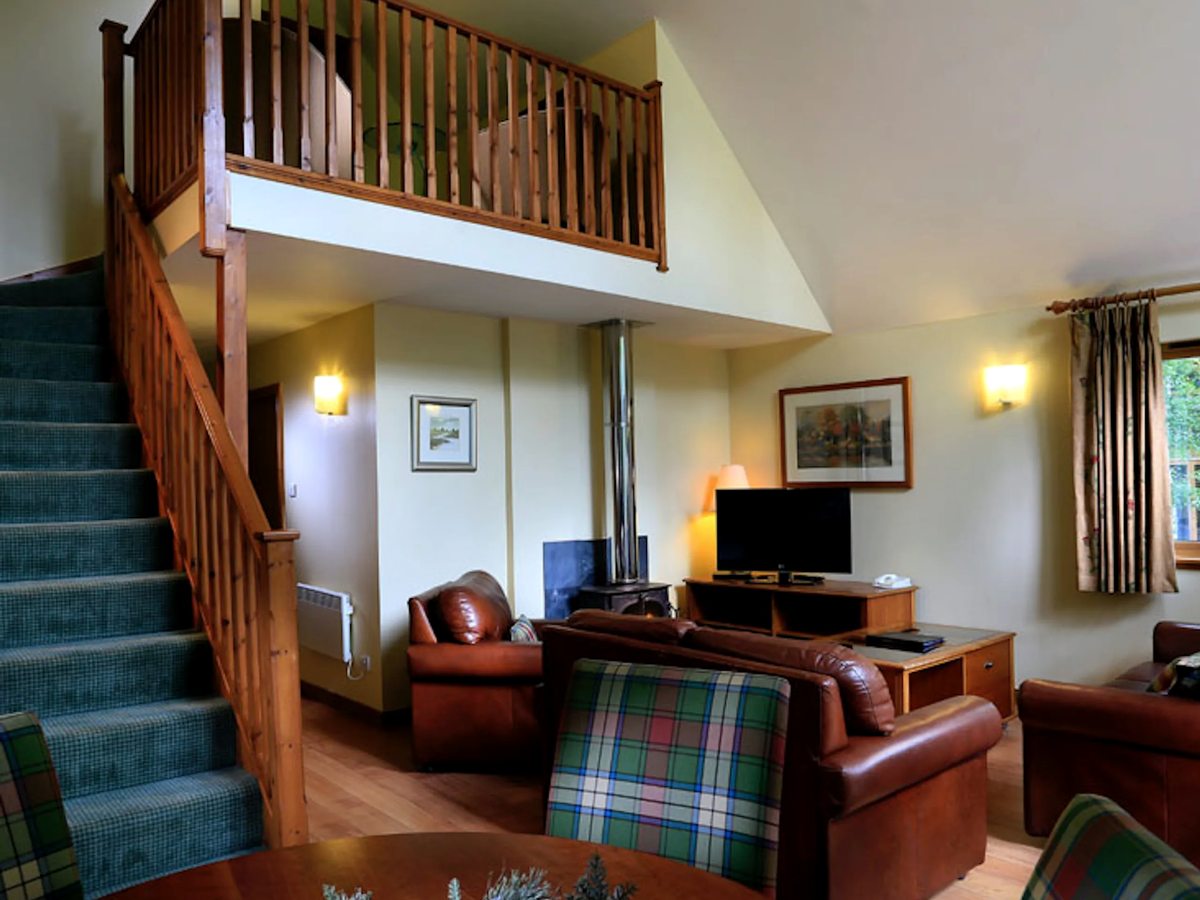 A lodge living room with tan leather sofas, tartan chairs, a television and a wood-burning stove, with a timber staircase up to a mezzanine.