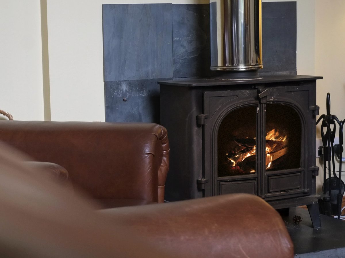 A black cast-iron wood-burning stove with a lit fire and a slate surround, with the arm of a brown leather sofa in front of it.
