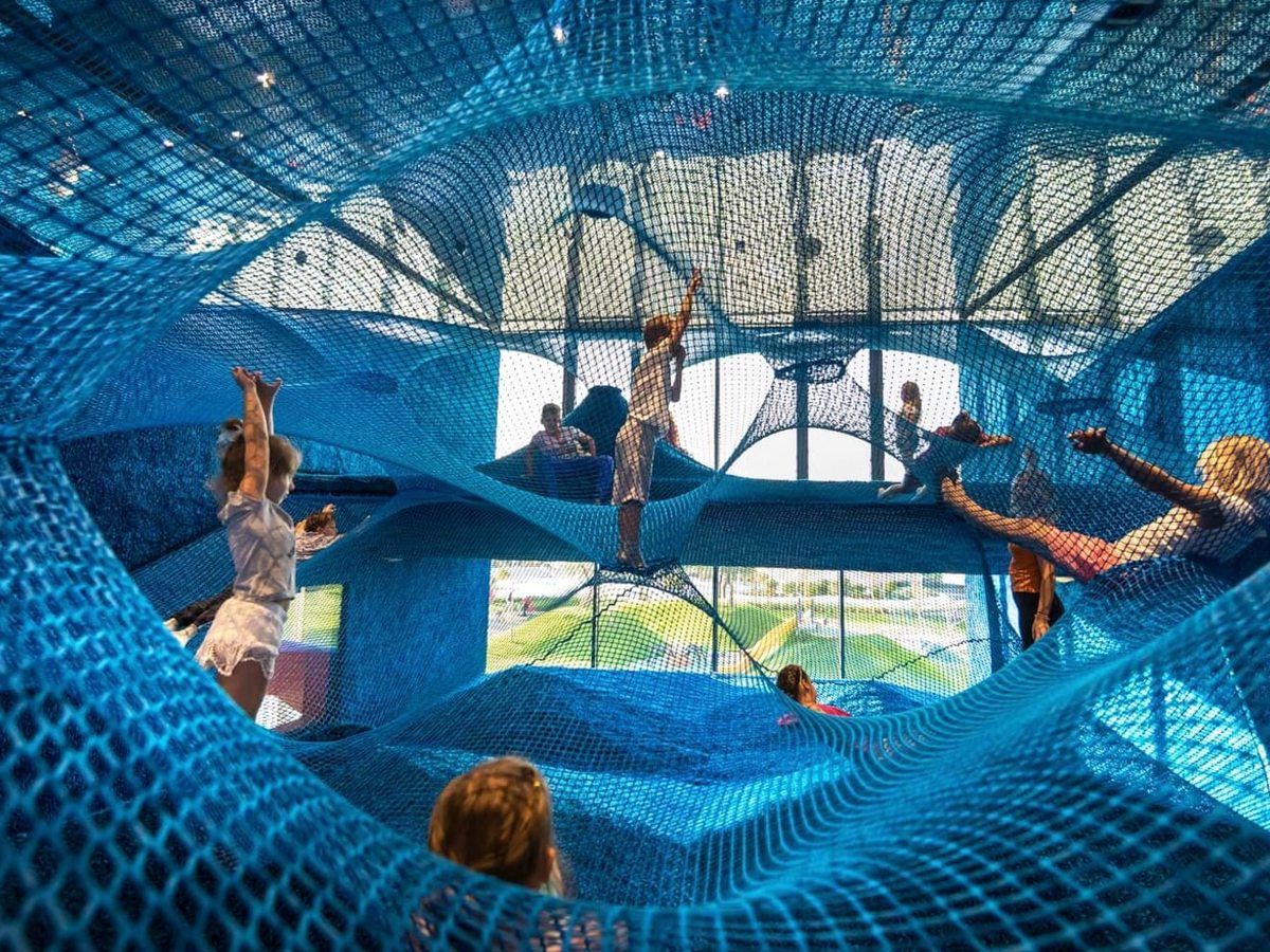 Children climbing and sitting inside a huge blue rope net suspended in curves across a bright atrium.