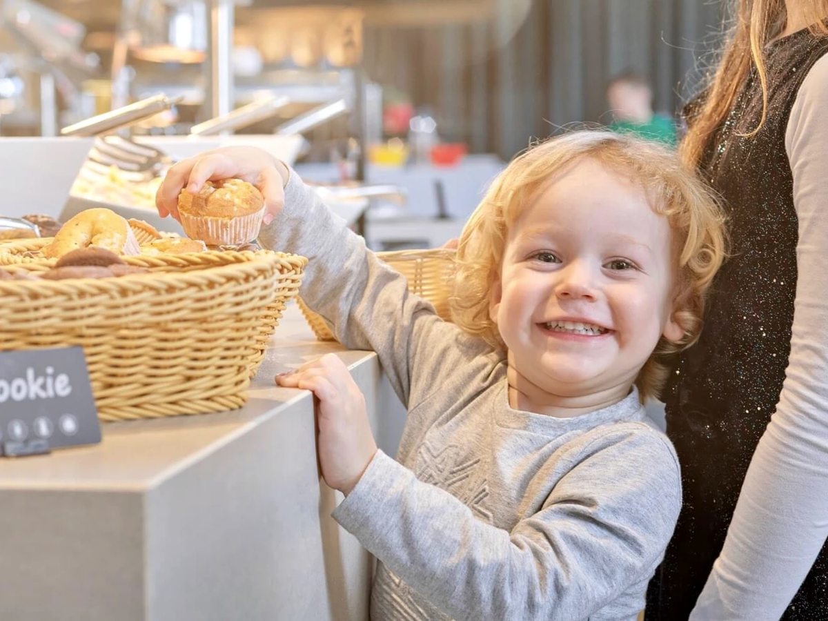 A toddler at a low buffet counter lifting a muffin from a wicker basket, with the buffet line behind.