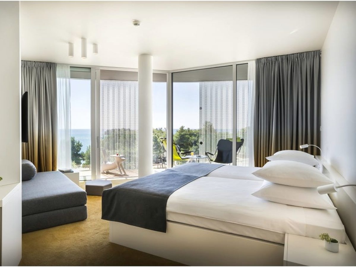 Corner hotel room with a double bed, a folded grey sofa bed and floor-to-ceiling glass onto a balcony with chairs, pines and sea beyond.