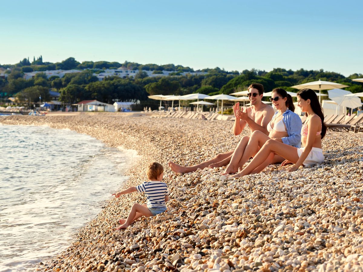 A family sitting on a white pebble beach at the water's edge with a toddler paddling, parasols and pines along the shore behind.