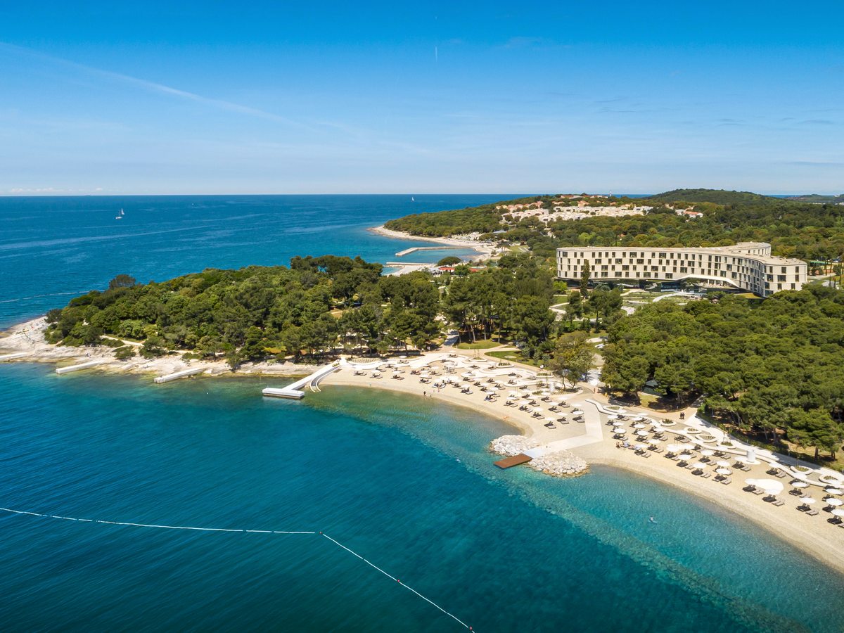 Aerial view of a long white four-storey hotel among pine woods on a headland, with a curving pebble beach lined with parasols and clear turquoise sea.