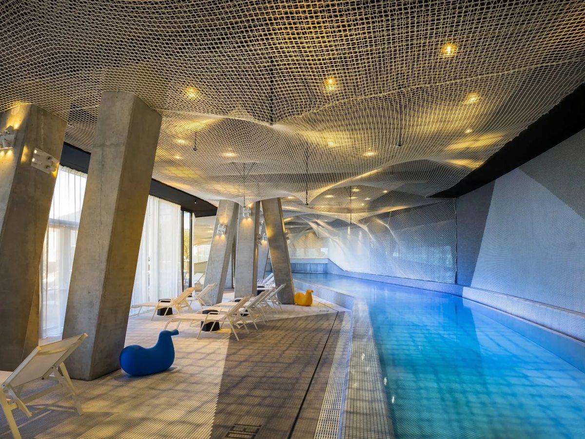 Long indoor pool lit blue, with angled concrete columns, white loungers and a rippling net canopy across the ceiling.